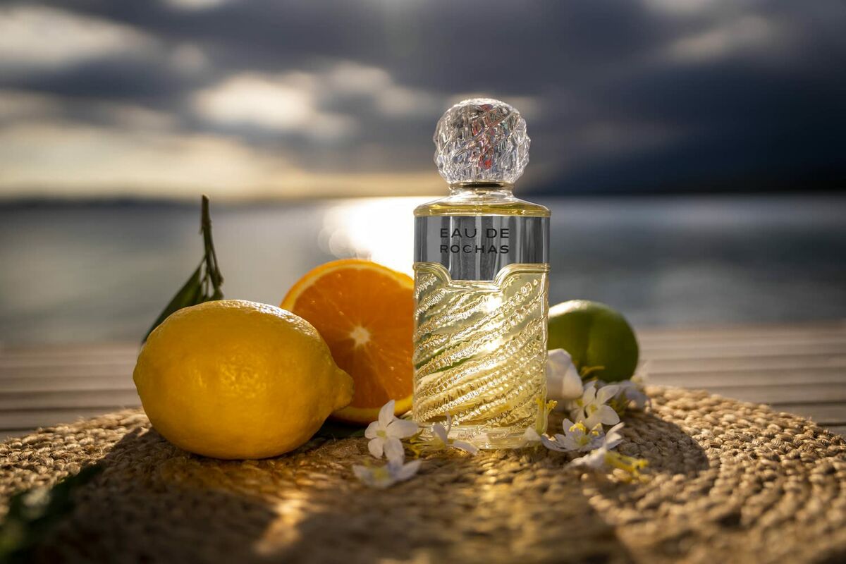 Bouteille de parfum Eau de Rochas avec des agrumes et des fleurs sur un fond naturel.