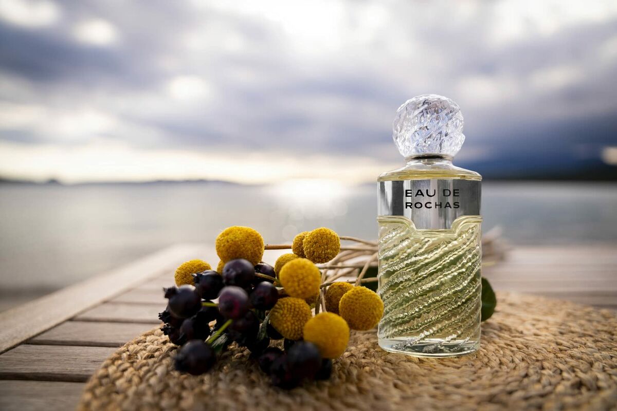 Bouteille de parfum Eau de Rochas sur une table près de l'eau, accompagnée de fleurs et de fruits.